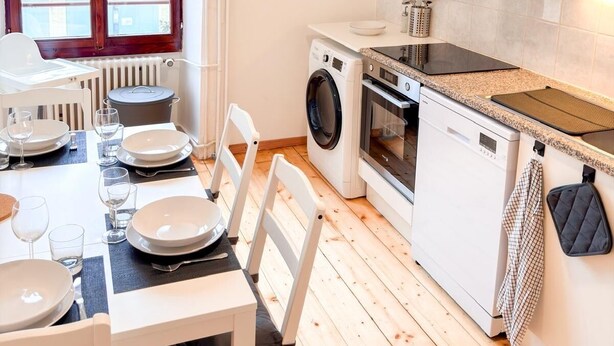 A compact kitchen with a washing machine, oven, and a dining area with chairs and a table set for two. - Central Geneva Apartment - Walk to Old Town & Lake (Genève)