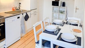 A modern kitchen with white cabinets, a dishwasher, and a dining area with white chairs and a table set for four. - Central Geneva Apartment - Walk to Old Town & Lake (Genève)