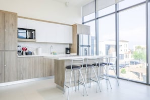 Eine moderne Küche mit weißen Schränken, einem Edelstahl-Kühlschrank, einer Mikrowelle und einer Kaffeemaschine. Drei Barhocker und ein großes Fenster bieten einen Blick ins Freie.