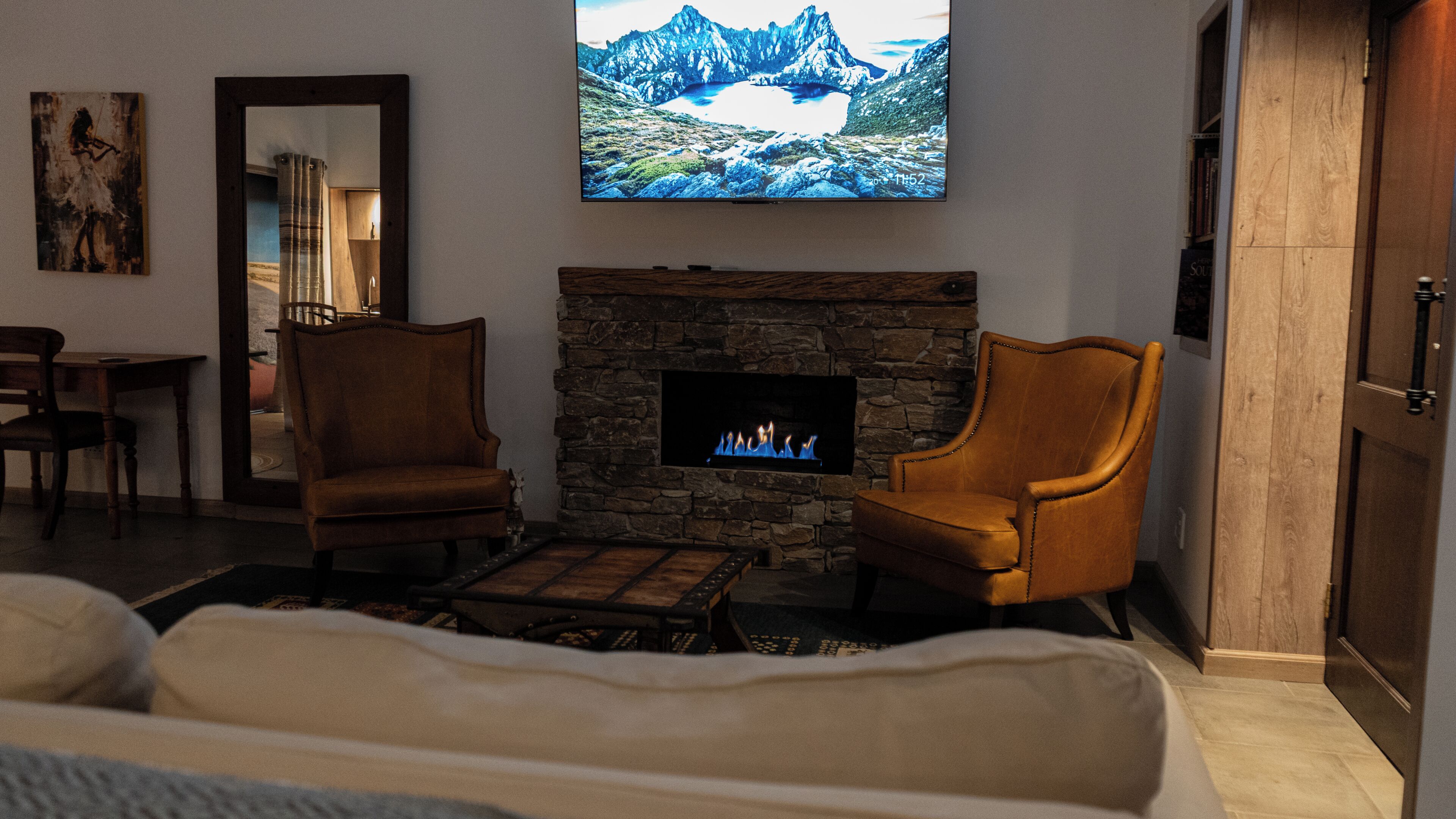 A living room with a stone fireplace, two armchairs, a sofa, and a flat-screen TV mounted above the fireplace displaying a mountain landscape.