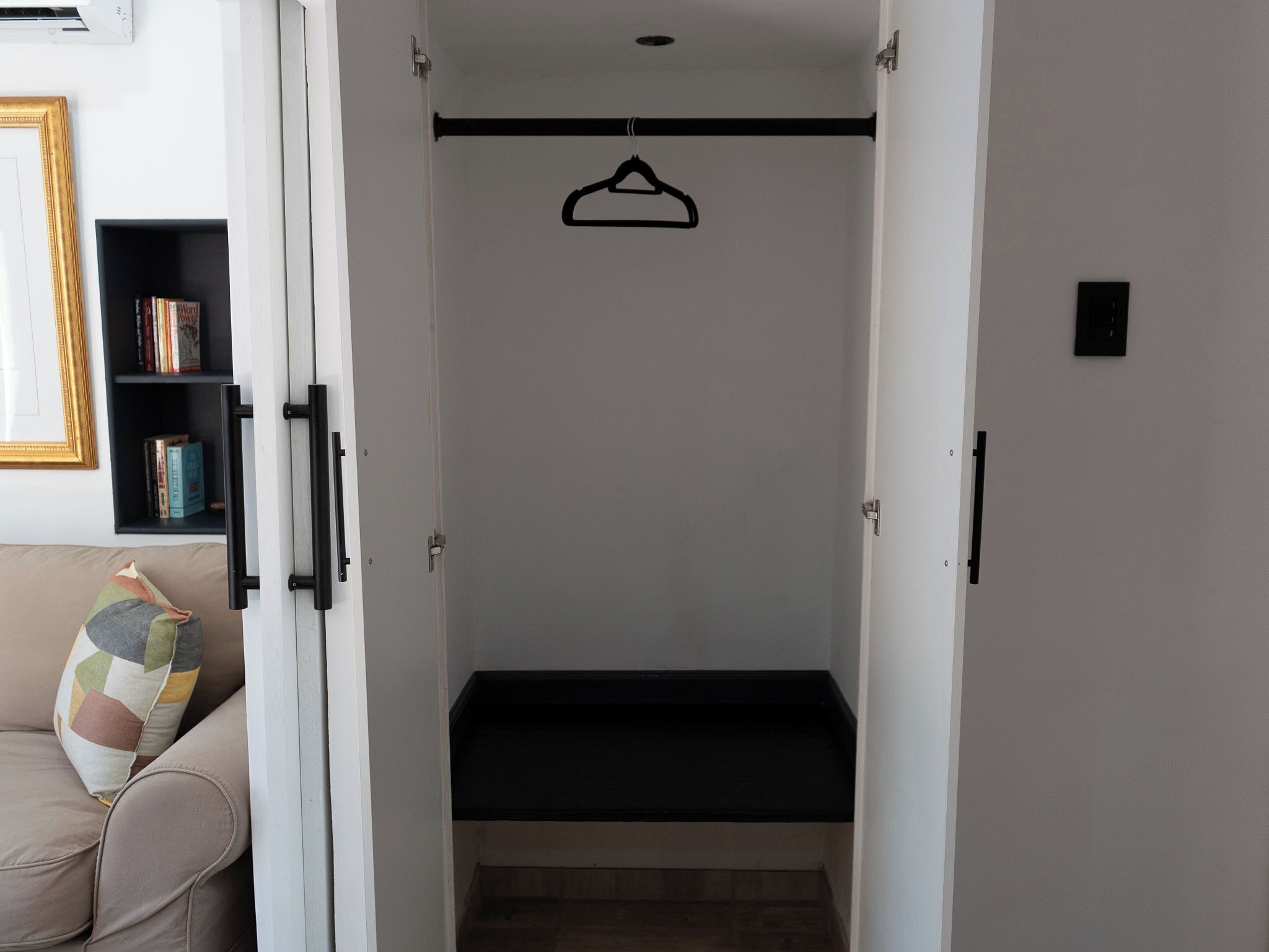 A room with a bed, a hanging clothes rack, a bookshelf, and a couch with a cushion.