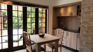 A dining area with a table and chairs, a window with a view of greenery, and a kitchen with wooden cabinets and a black countertop. - The Lombard Manor (Pretoria)