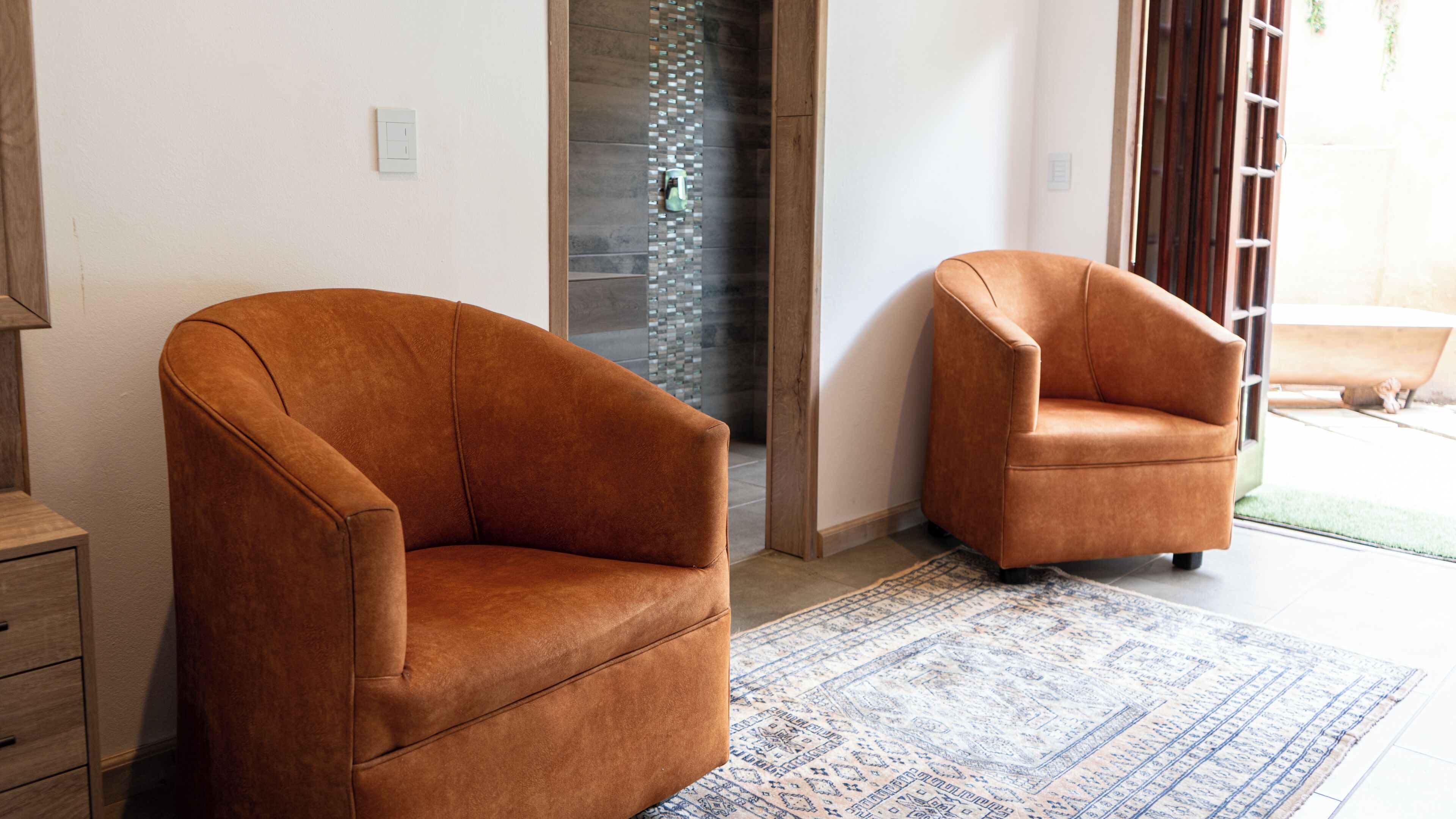 A room with two brown armchairs, a patterned rug, a wooden cabinet, and a glass door leading to an outdoor area.
