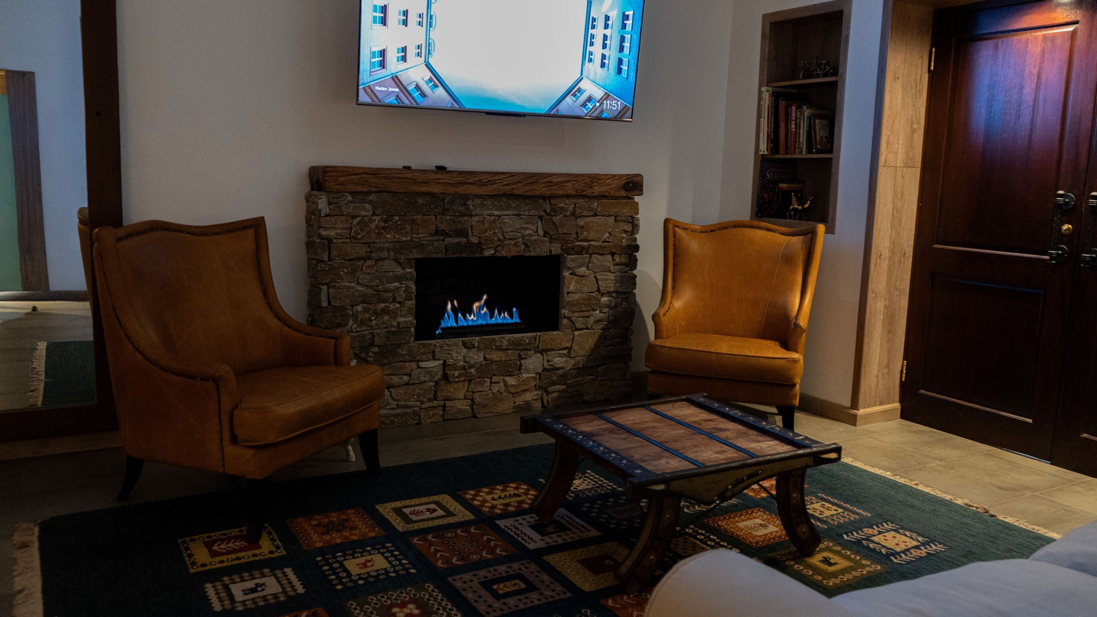 A living room with a stone fireplace, two leather armchairs, a coffee table, and a flat-screen TV mounted on the wall.