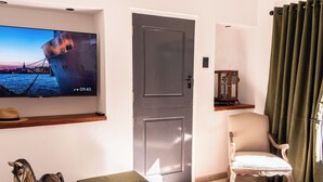 Un salón con una televisión mostrando una escena de atardecer, una puerta gris, un caballo balancín, un reposapiés verde, un sillón beige y una alfombra tejida con colores vivos.