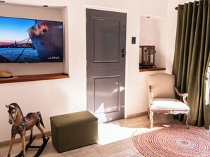 A living room with a television displaying a sunset scene, a grey door, a rocking horse, a green ottoman, a beige armchair, and a colorful woven rug.