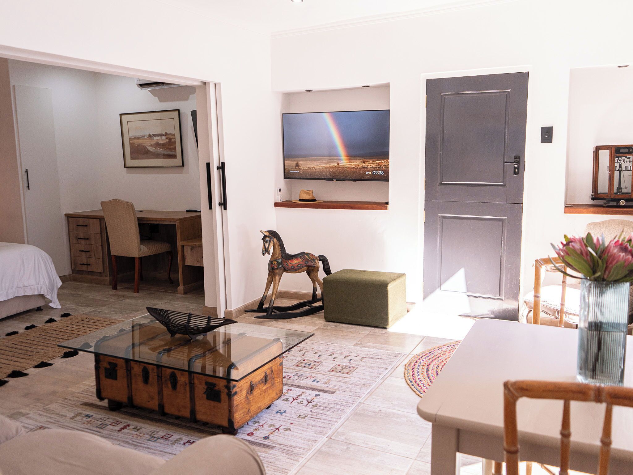 Een goed verlichte kamer met een bed, een bureau, een televisie, een deur en een kleine houten paard decoratie.
