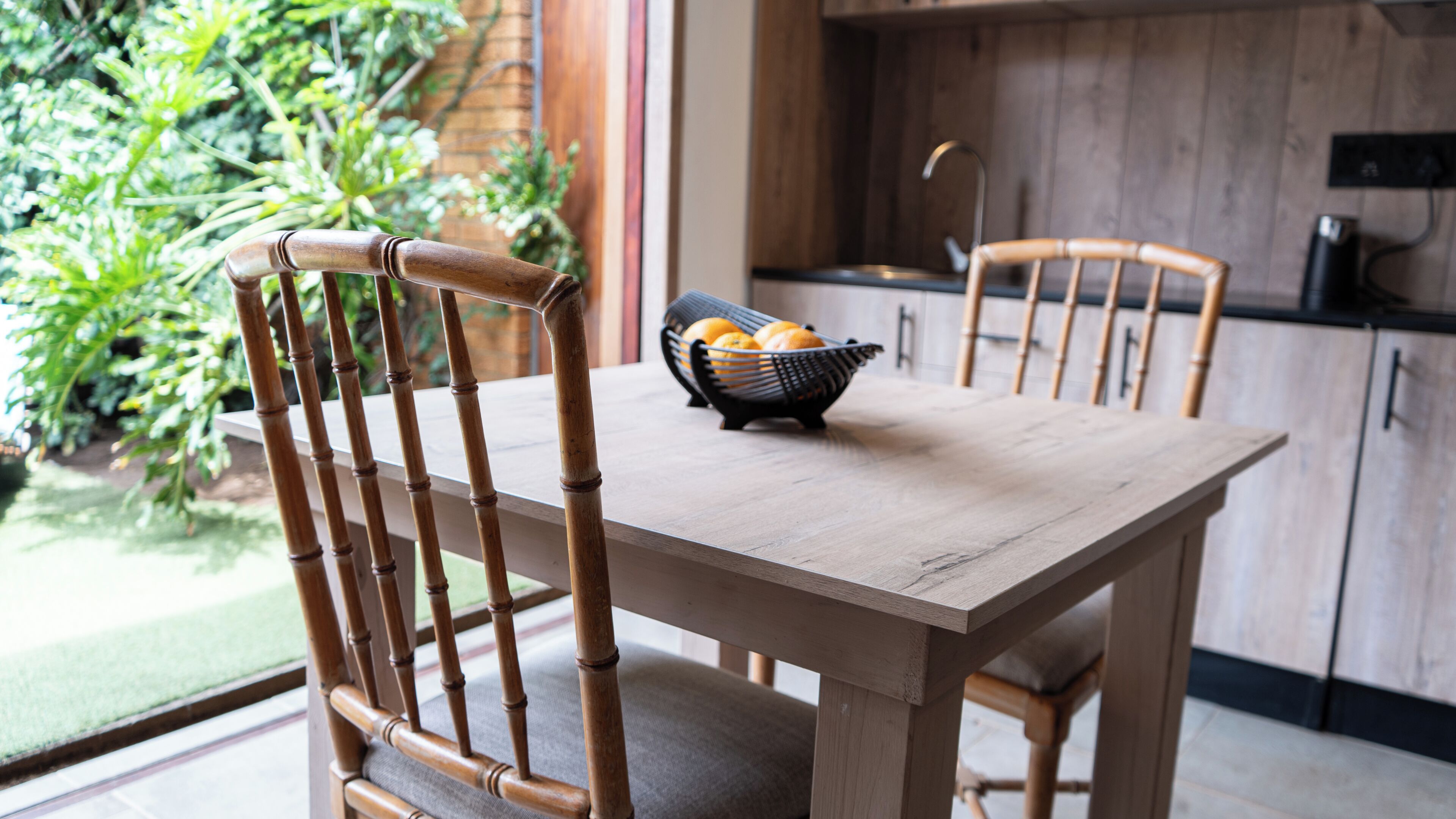 Una mesa de madera con sillas, una cesta de naranjas y una cocina con armarios y un lavamanos.