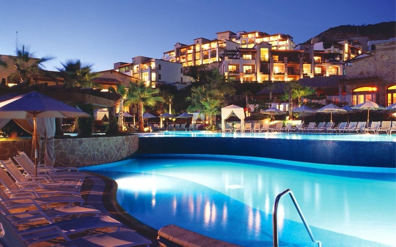 A swimming pool with lounge chairs and umbrellas, surrounded by a resort building with multiple balconies.