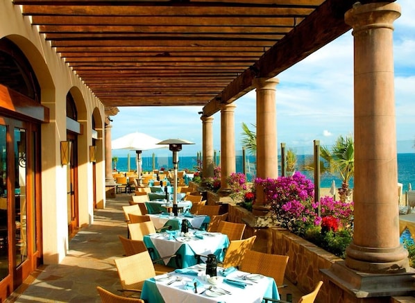 A covered outdoor dining area with tables and chairs, overlooking a beach.