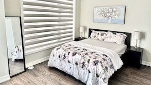 A bedroom with a bed, two bedside tables, a nightstand, a mirror, and a window with blinds. - Modern 3 bed, 2 bath townhouse. (Vaughan)