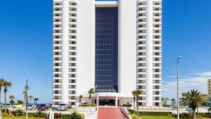 Um hotel alto e branco, com fachada central de vidro, várias varandas e um caminho de entrada vermelho.