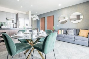 Een moderne keuken met roestvrijstalen apparatuur, een eethoek met een glazen tafel en groen gestoffeerde stoelen, en een grijze bank met gekleurde kussens.