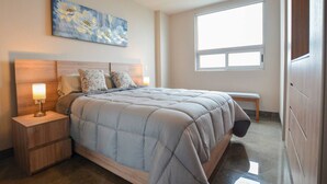 A bedroom with a large bed, a nightstand with a lamp, a bench, and a window. - ¡Moderno y de lujo! - Fundidora, Cintermex, arena (Monterrey)