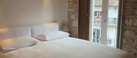 Un dormitorio con una cama grande, ropa de cama blanca, una pared de piedra y un balcón con barandilla de metal.