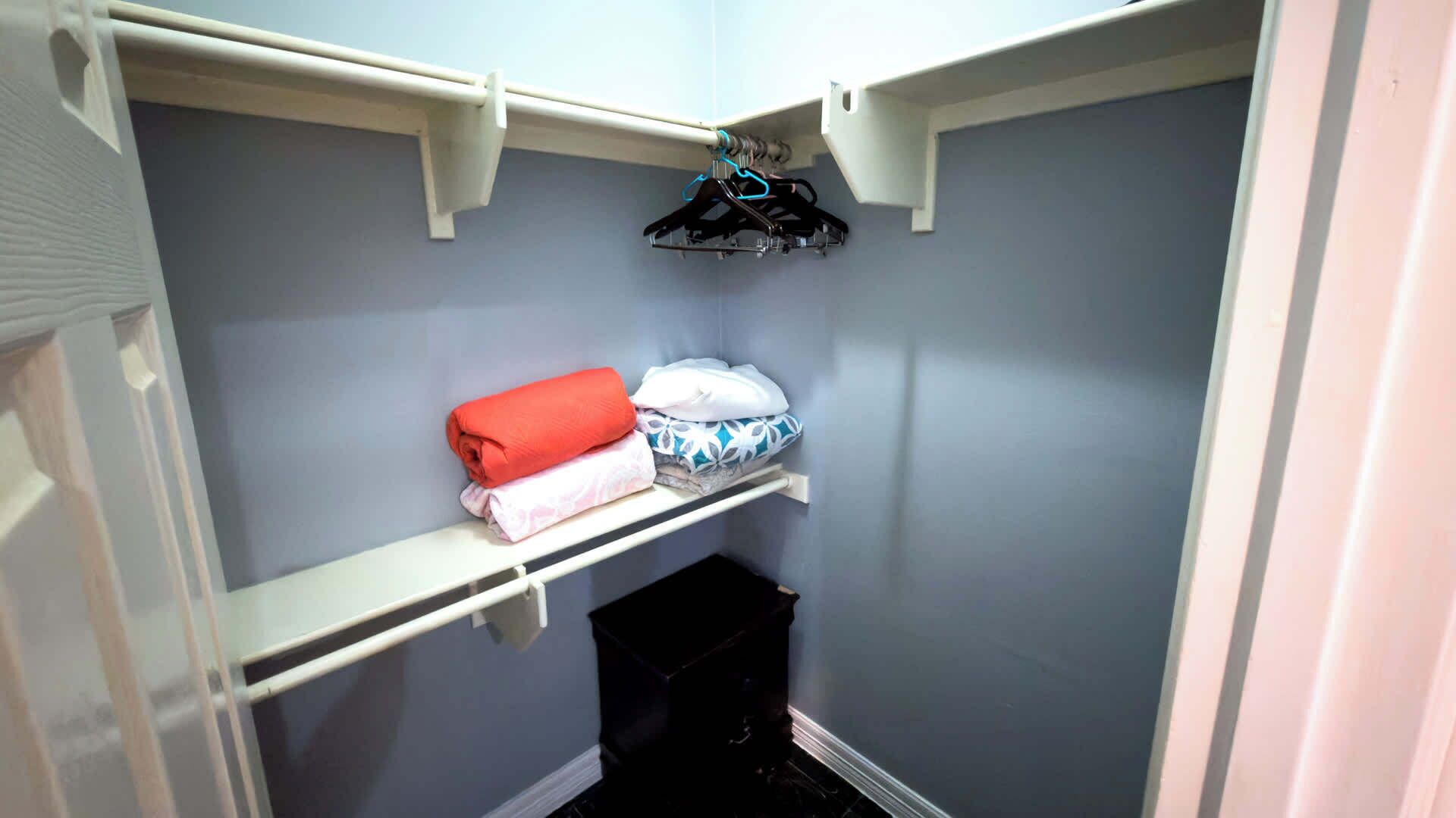 Een kleine kast met een wit plankje, waarop opgevouwen kleding ligt, een zwarte prullenbak en een blauwe kapstok aan een witte stang.