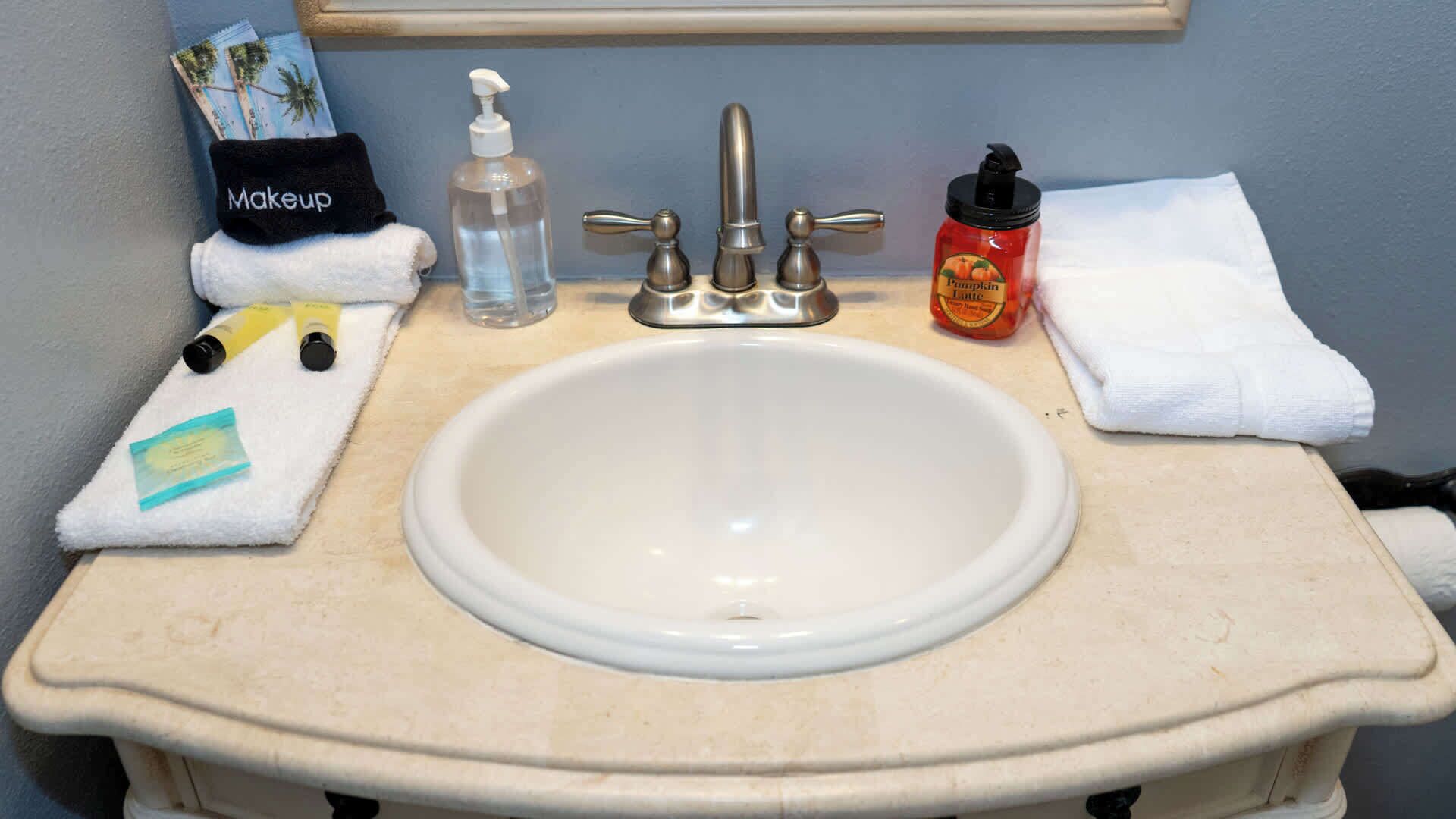 A bathroom sink with a mirror above it, a towel, a bottle of hand soap, a bottle of hand sanitizer, and a bottle of pumpkin spice body wash.