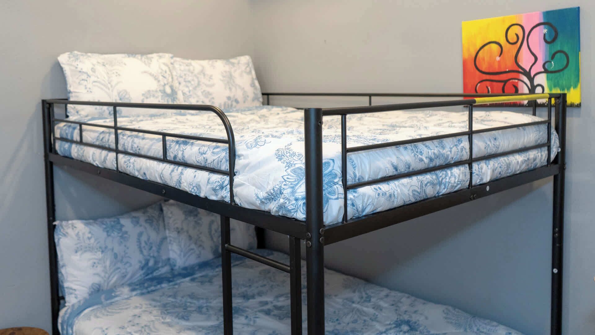 Uma cama beliche com colchão e almofadas estampadas, um quadro abstrato colorido na parede e estrutura de metal.