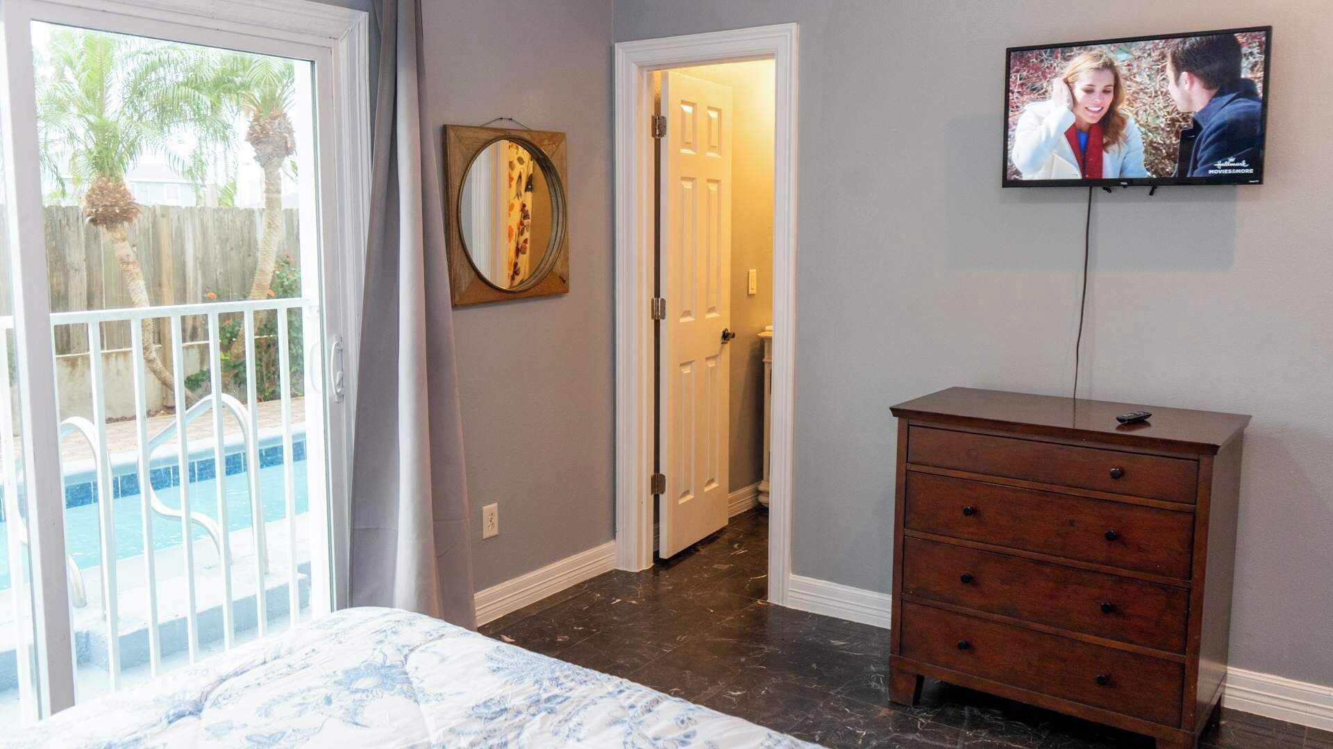 A bedroom with a bed, a wooden dresser, a TV on the wall, and a door leading to another room.