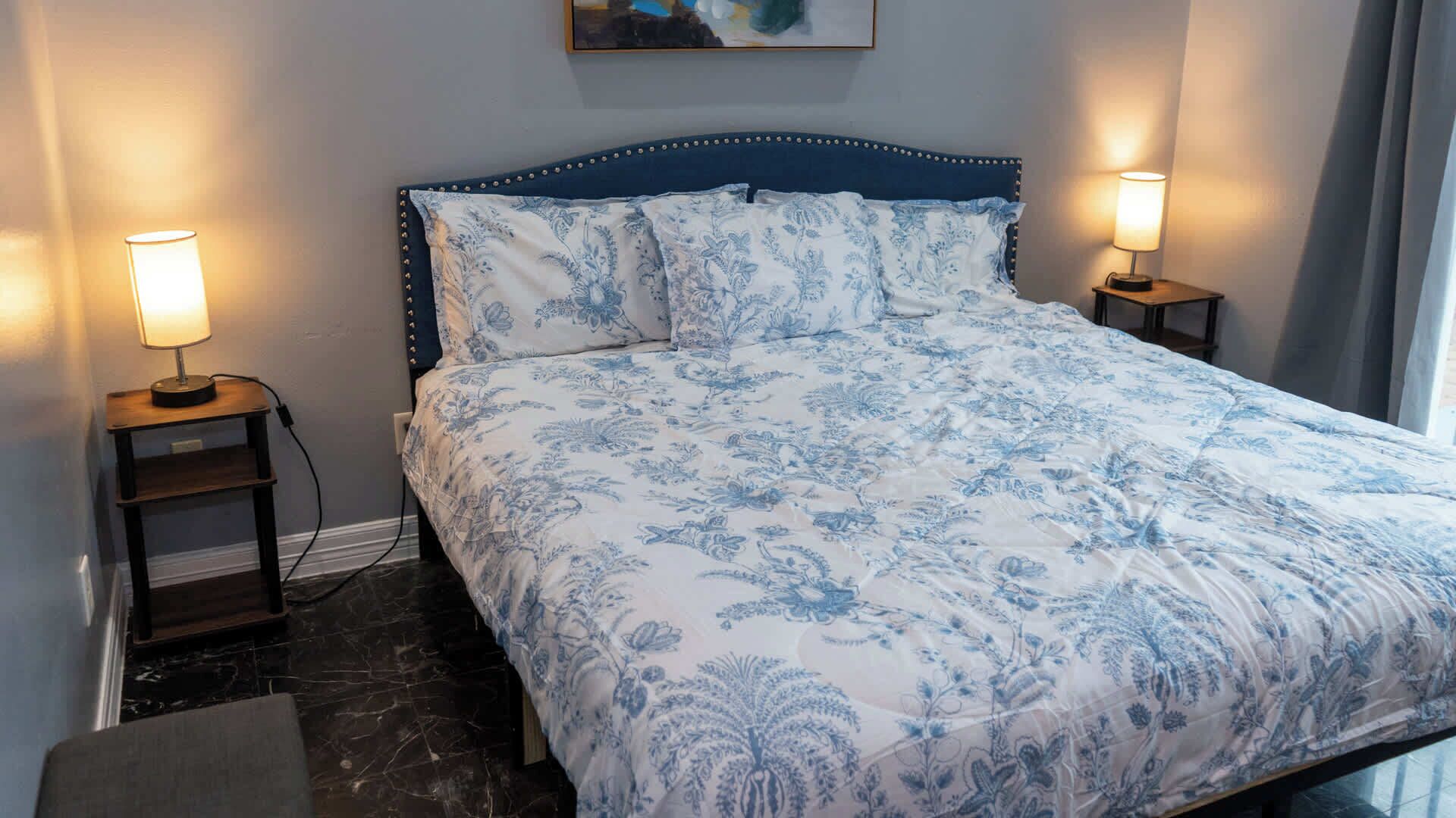 A neatly made bed with a floral pattern, flanked by two bedside tables with lamps, in a room with a marble floor and a painting on the wall.