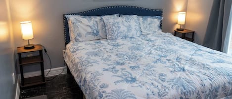 A neatly made bed with a floral pattern, flanked by two bedside tables with lamps, in a room with a marble floor and a painting on the wall.