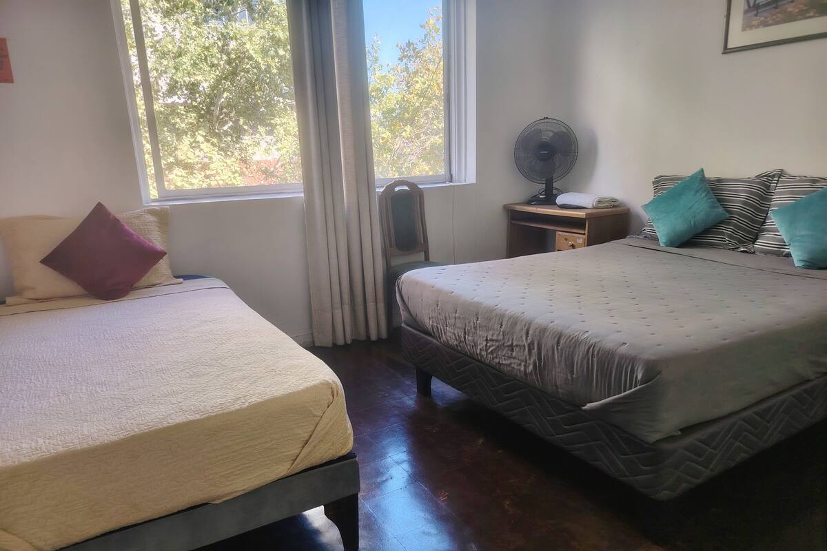 A room with two beds, a window with curtains, a standing fan, and a desk with a chair.