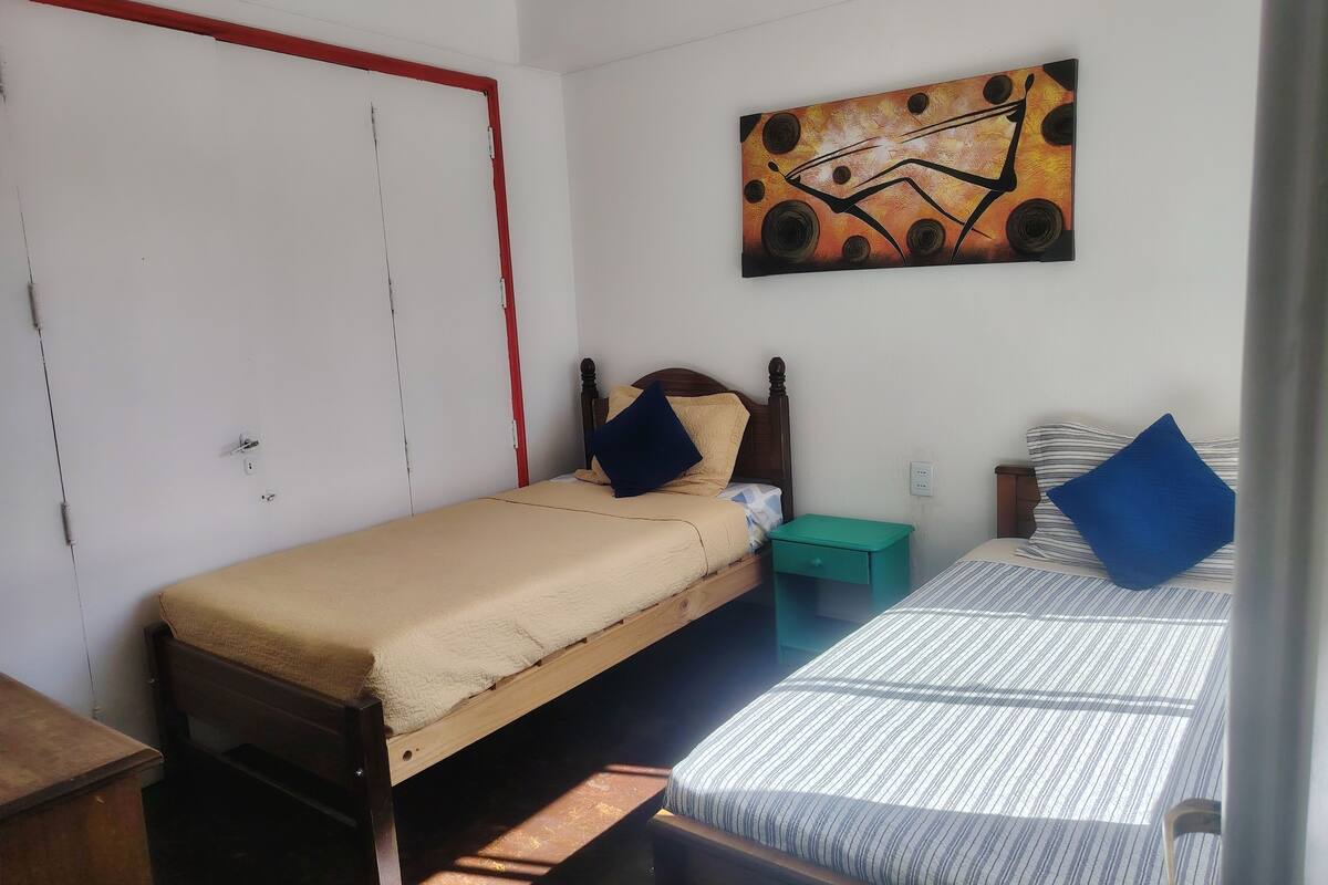 A small, simple room with two beds, a wooden wardrobe, a green nightstand, and a wall painting.