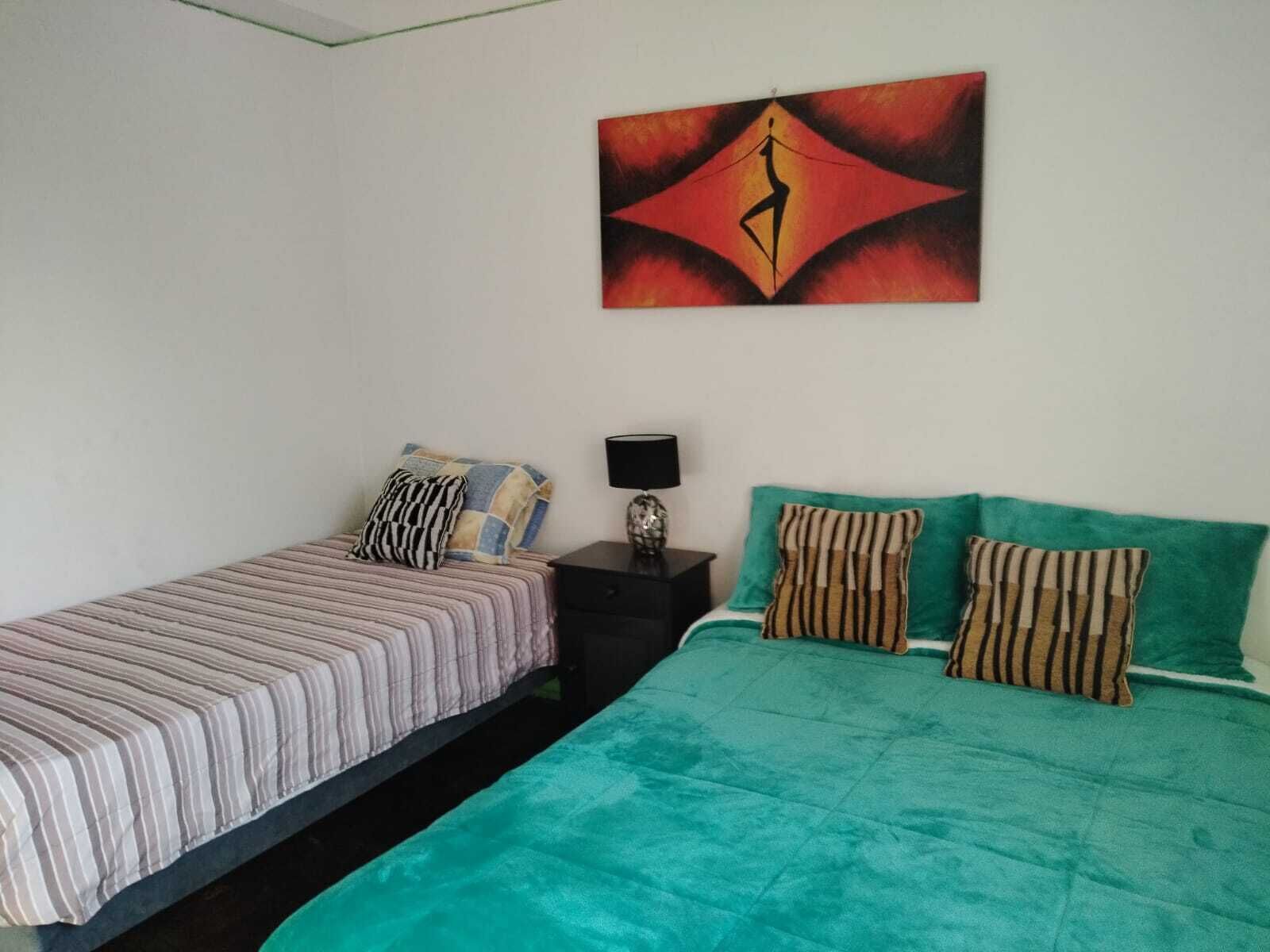 Un dormitorio con dos camas, una con ropa de cama a rayas y la otra con ropa de cama verde. Hay una mesita de noche con una lámpara y un objeto decorativo. Un cuadro cuelga de la pared sobre las camas.