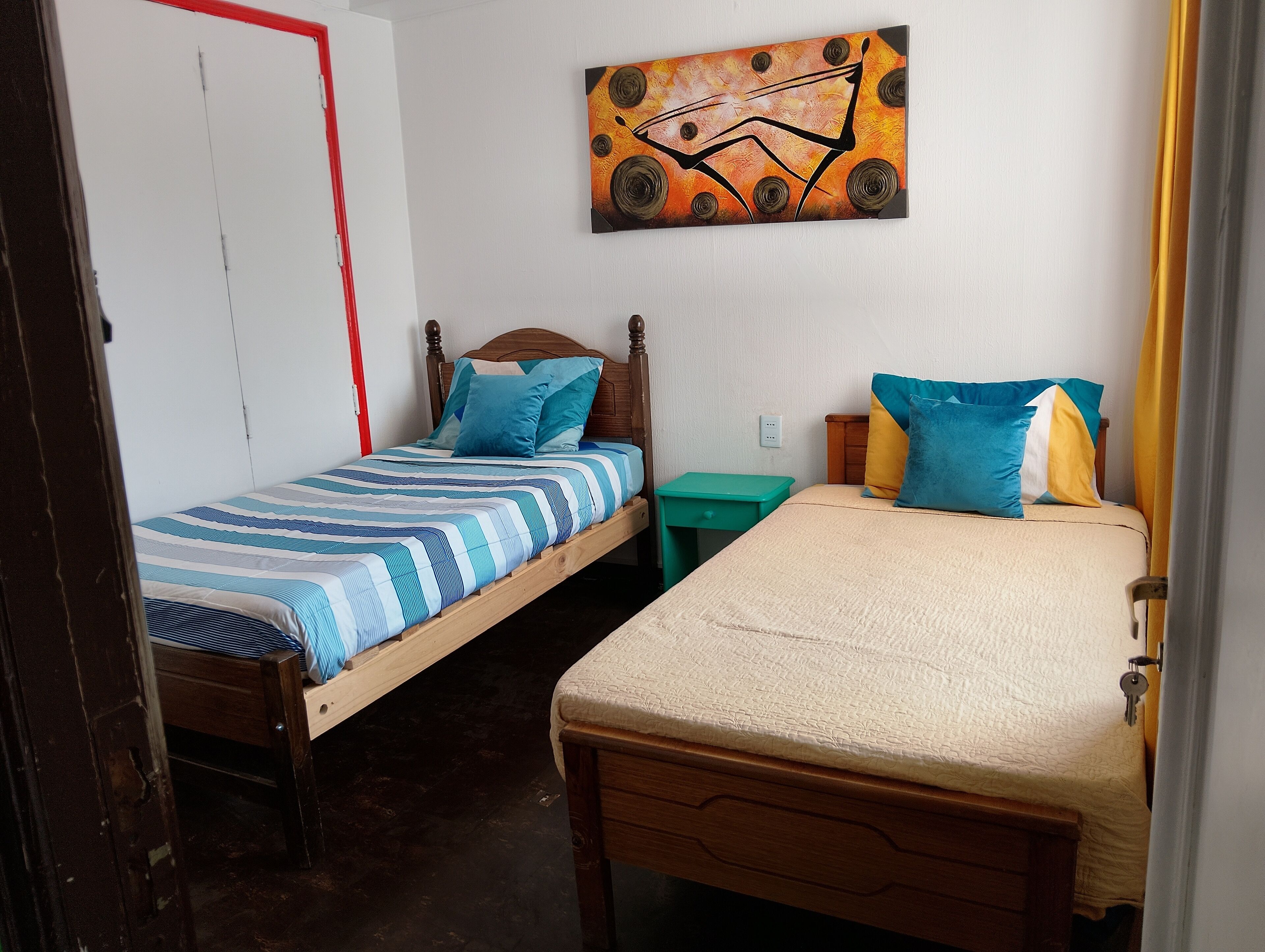 A room with two wooden beds, one with a striped blue and white bedspread and the other with a beige bedspread. There is a green nightstand with a lamp, a painting on the wall, and a white door with a red handle.
