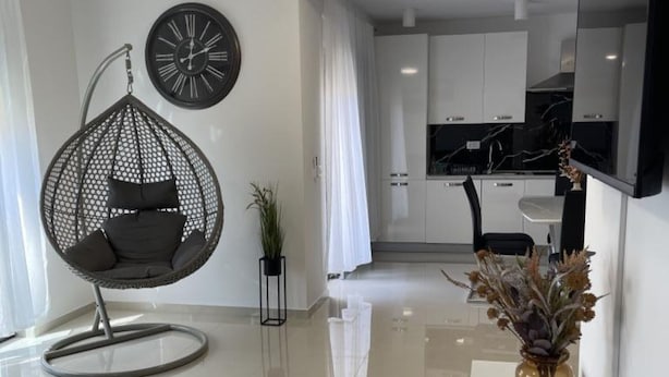 Ein modernes Wohnzimmer mit einem Hängekorbstuhl, einer Uhr, einem Blumentopf und einer Küche im Hintergrund.