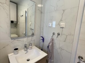 A bathroom with a marble wall, a sink with a cup and bottle, a towel hanging, and a shower area with a blue mop. - Apartment Rogoznica-Two Bedroom Apartment 1 (Rogoznica)