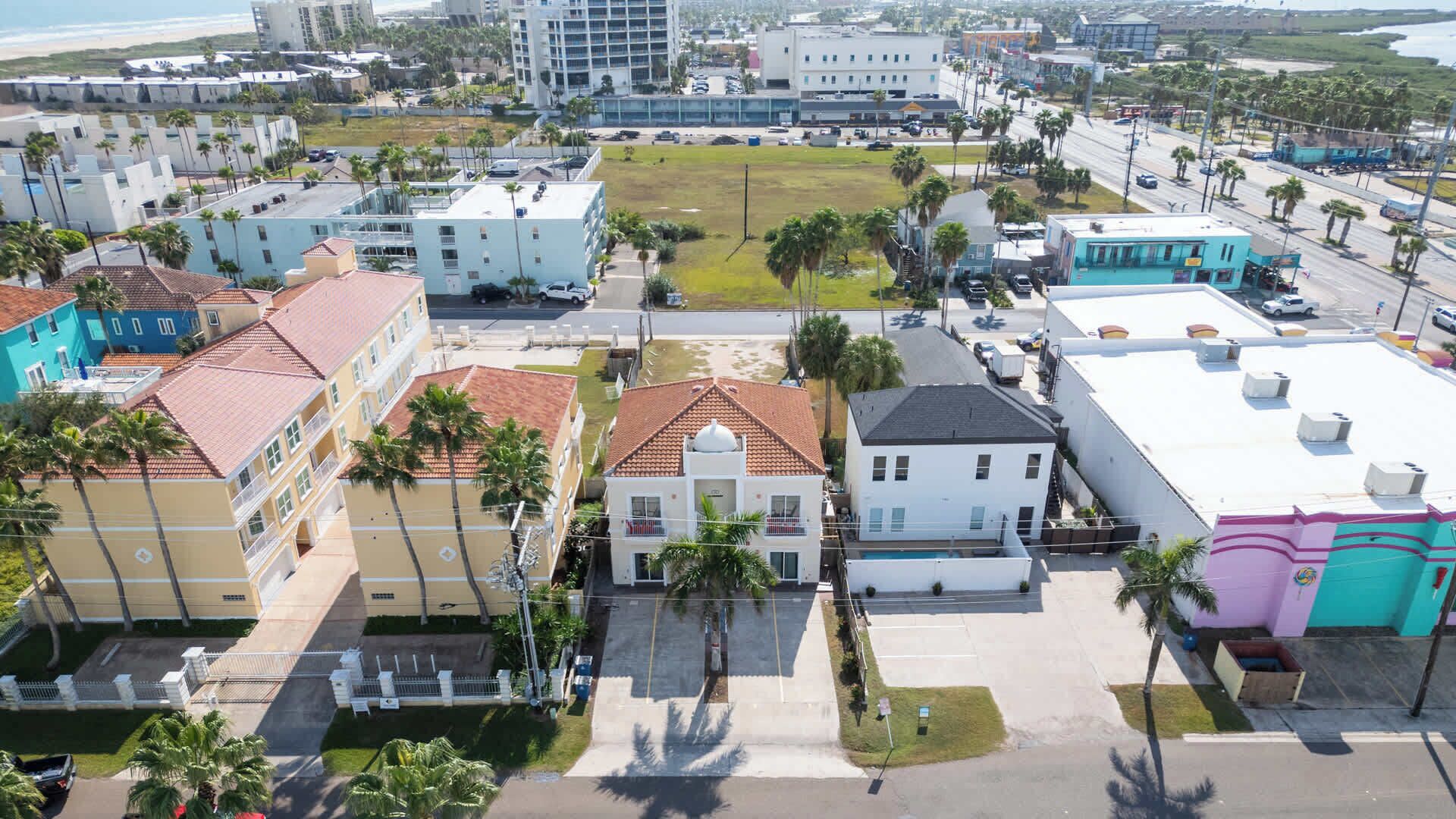 Vista aérea de una zona urbana costera con varios edificios, incluyendo un edificio blanco destacado con techo rojo y un edificio azul con techo blanco.