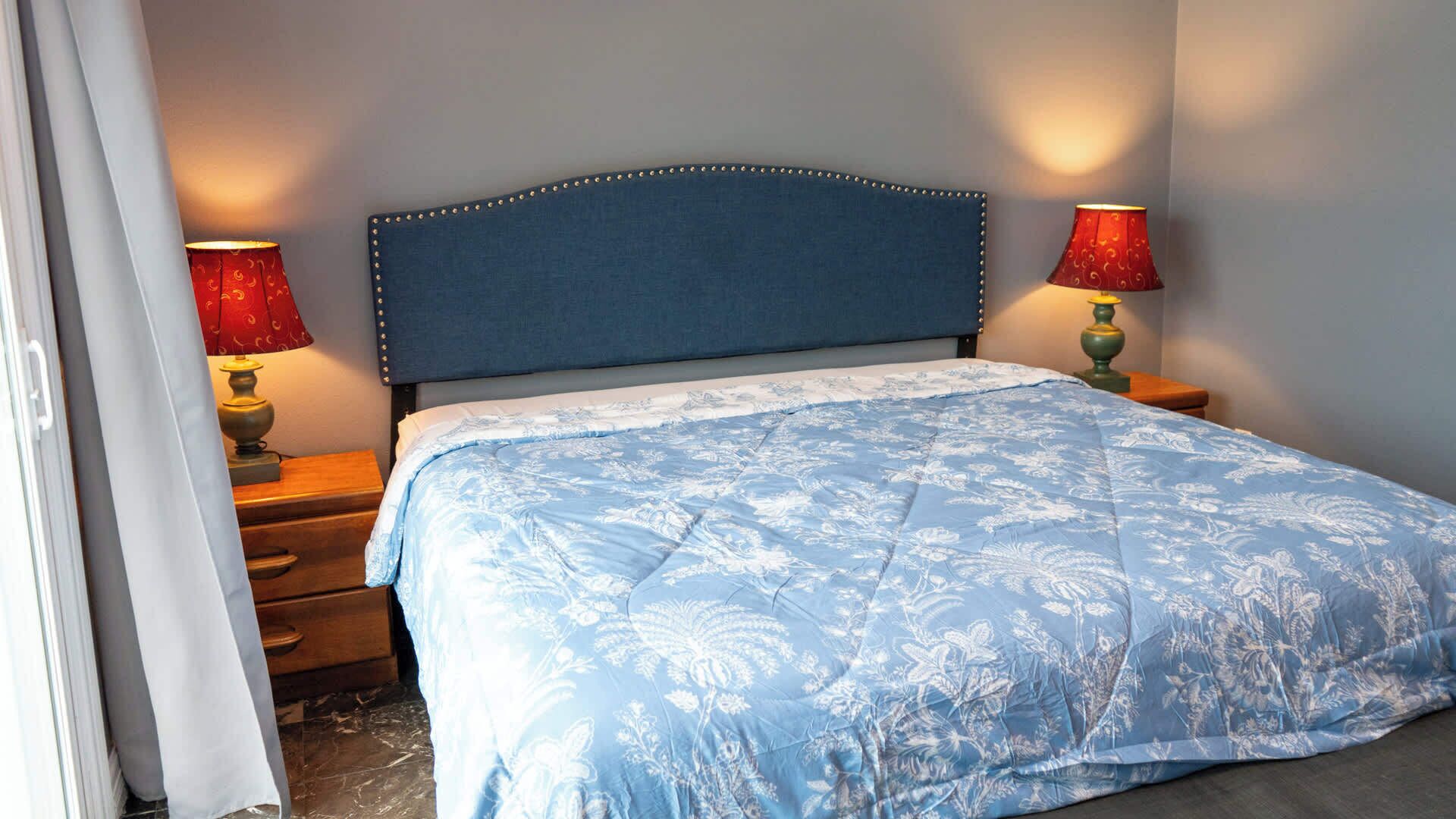 A bedroom with a blue headboard, a floral bedspread, two bedside lamps, and a wooden nightstand.