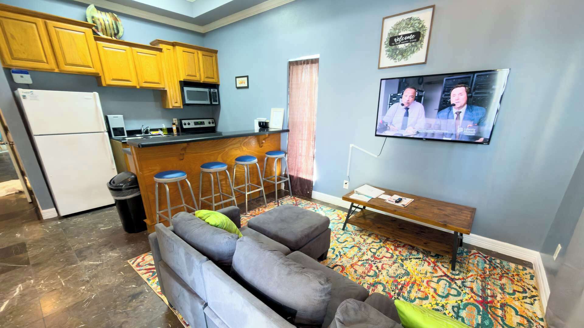 A compact living space with a kitchenette, a gray sofa with a green pillow, a colorful rug, and a TV mounted on the wall.