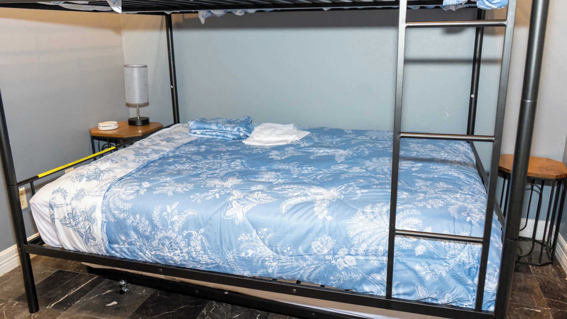 Una cama litera con una colcha azul estampada con flores, una mesita de noche con lámpara y un taburete de madera.