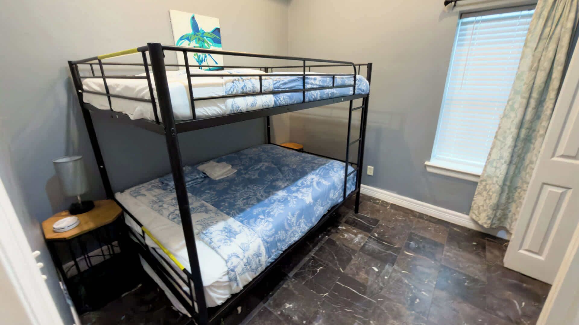 A bunk bed room with a marble floor, a small bedside table, and a window with curtains.