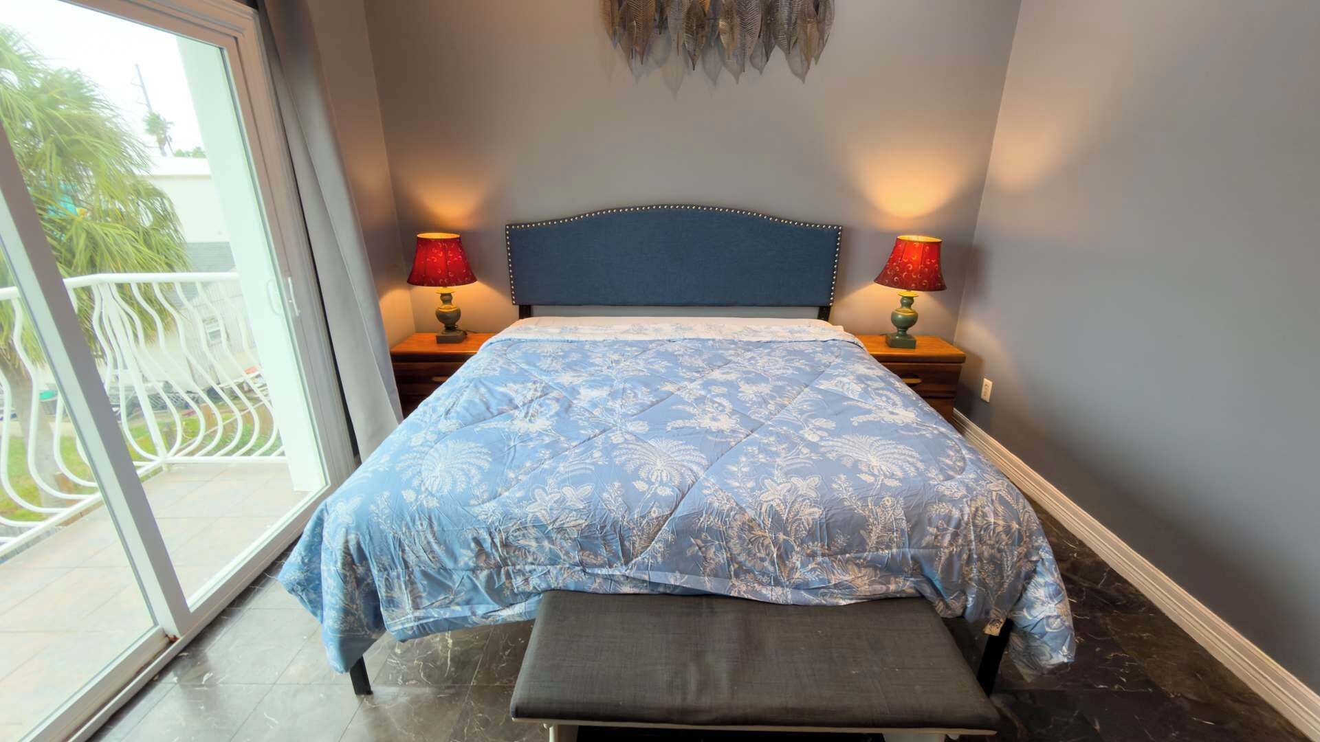 A bedroom with a bed, two bedside lamps, a bench, and a view of a balcony with a railing.