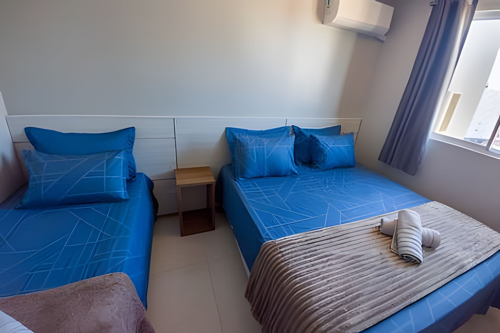 Un dormitorio con dos camas, una mesita de noche de madera, una ventana con cortinas y una toalla sobre la cama.
