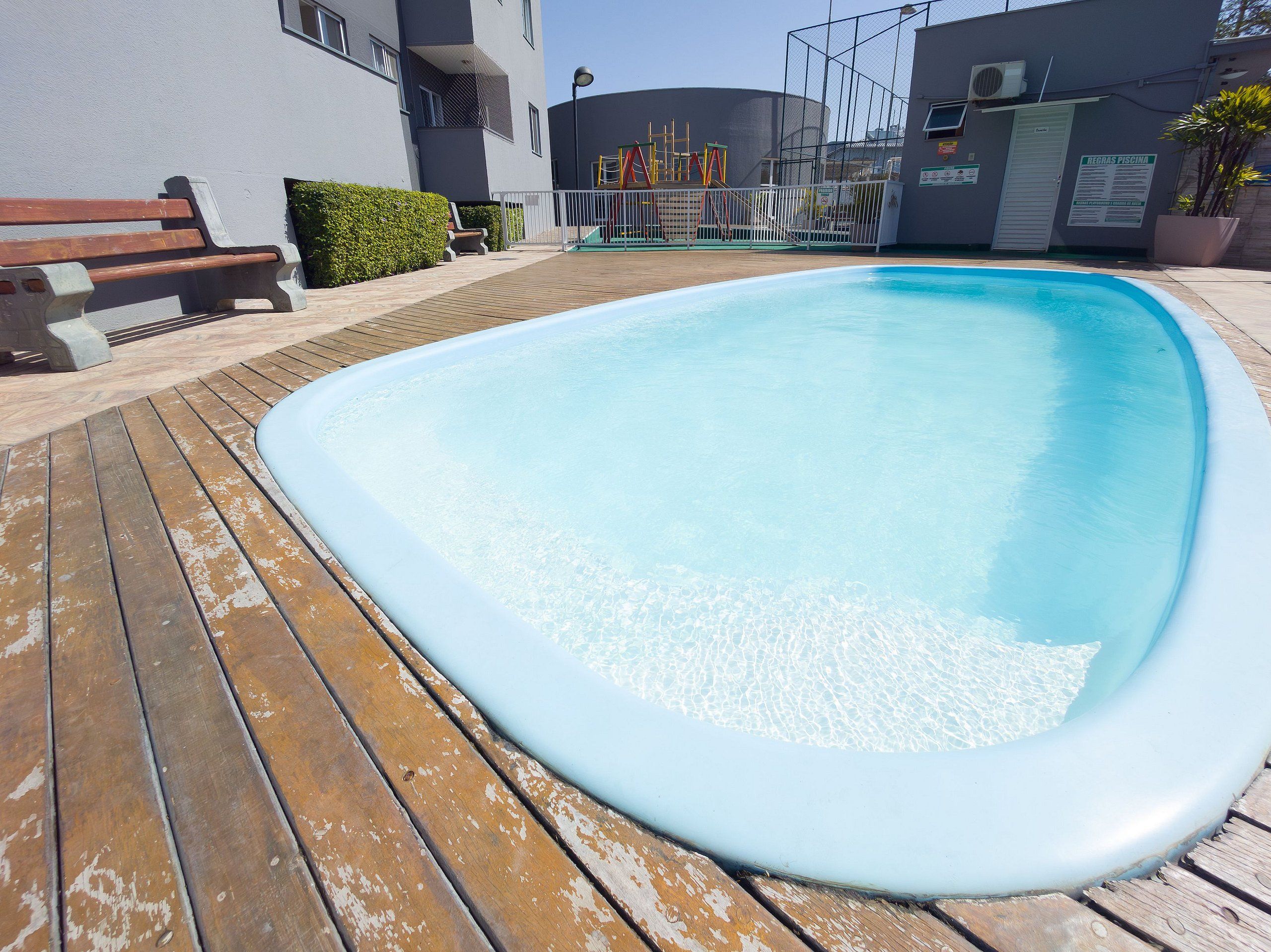 Una piscina con cubierta de madera, una banca y un parque infantil al fondo.