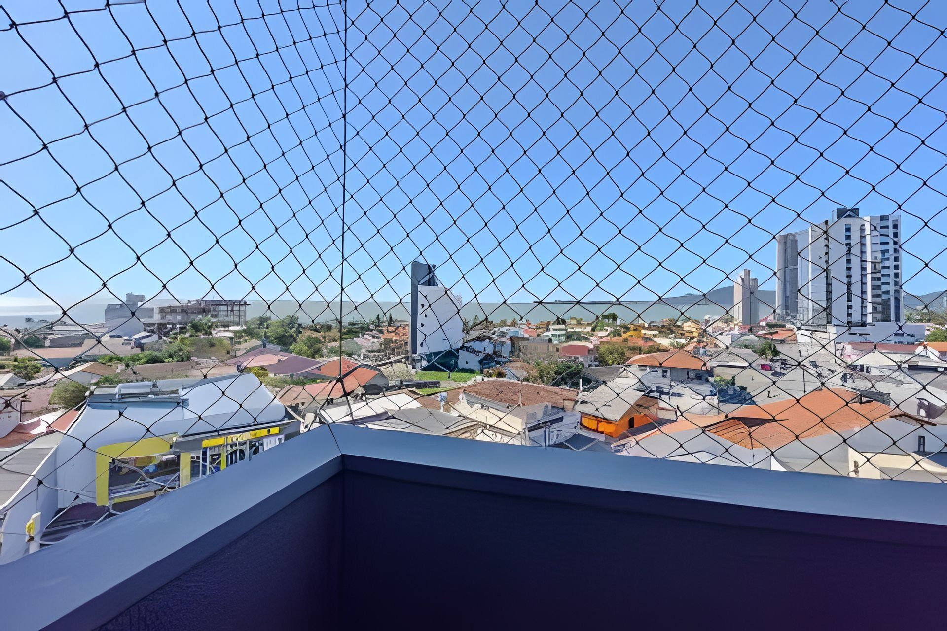 A cityscape through a chain-link fence with buildings, a clear sky, and a distant view of the sea.