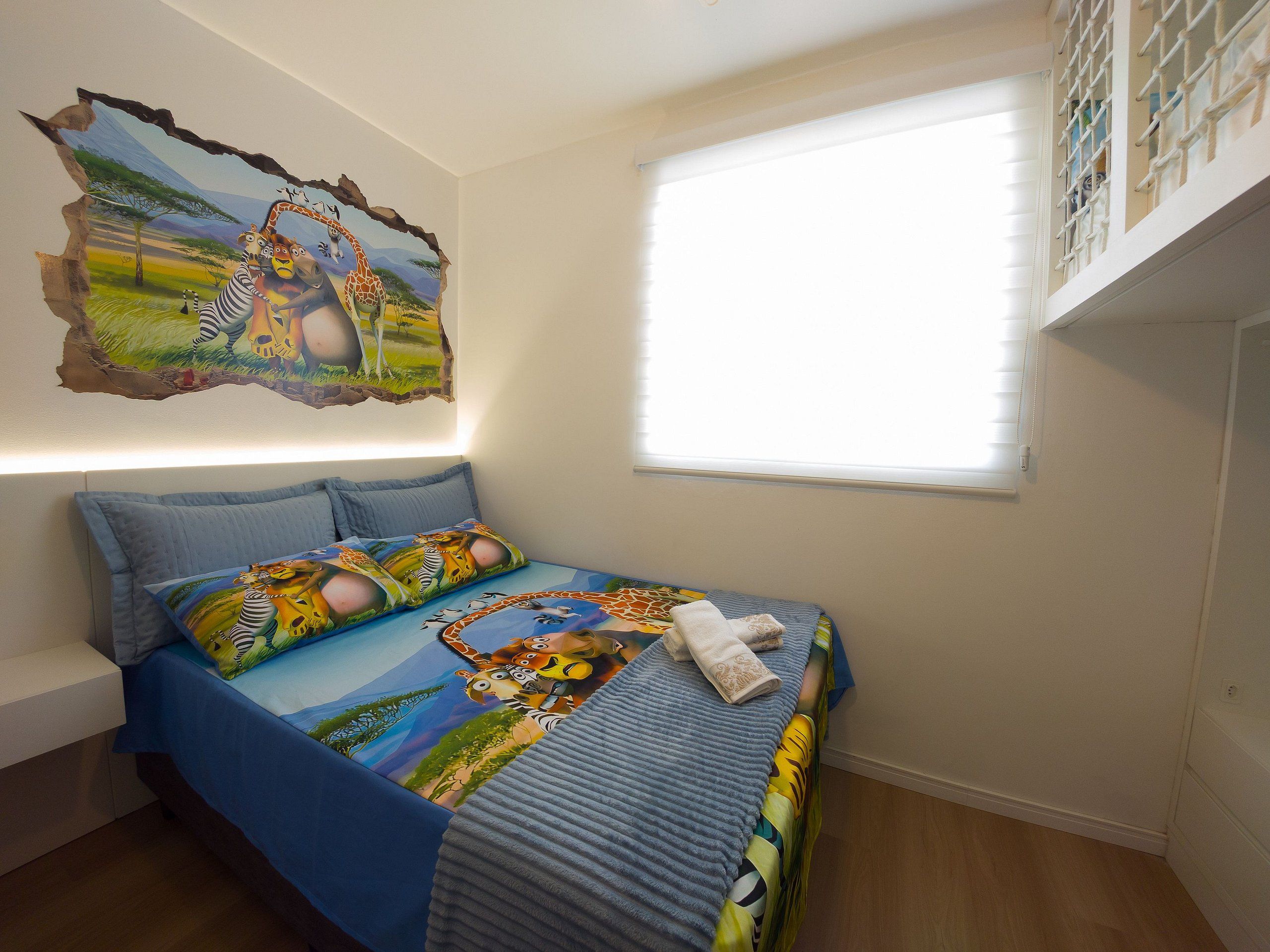A bedroom with a bed featuring a colorful animal-themed bedspread, a wall mural of animals, a window with blinds, and a white bench.
