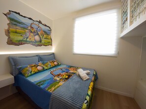 A bedroom with a bed featuring a colorful animal-themed bedspread, a wall mural of animals, a window with blinds, and a white bench. - Curta Praia do Quilombo - Lunata (Penha)