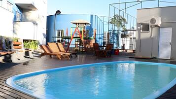 A swimming pool with lounge chairs, a children's play structure, and a large window.