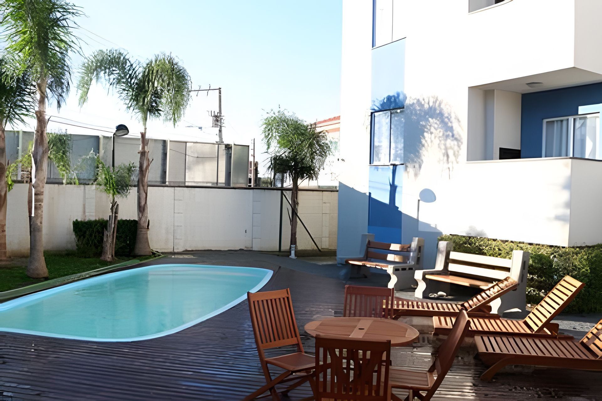 A swimming pool with wooden deck chairs and a table, surrounded by a white building with blue accents.