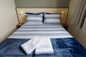 A neatly made bed with striped bedding, two pillows, and a folded towel on a wooden nightstand. - Curta Praia do Quilombo - Lunata (Penha)