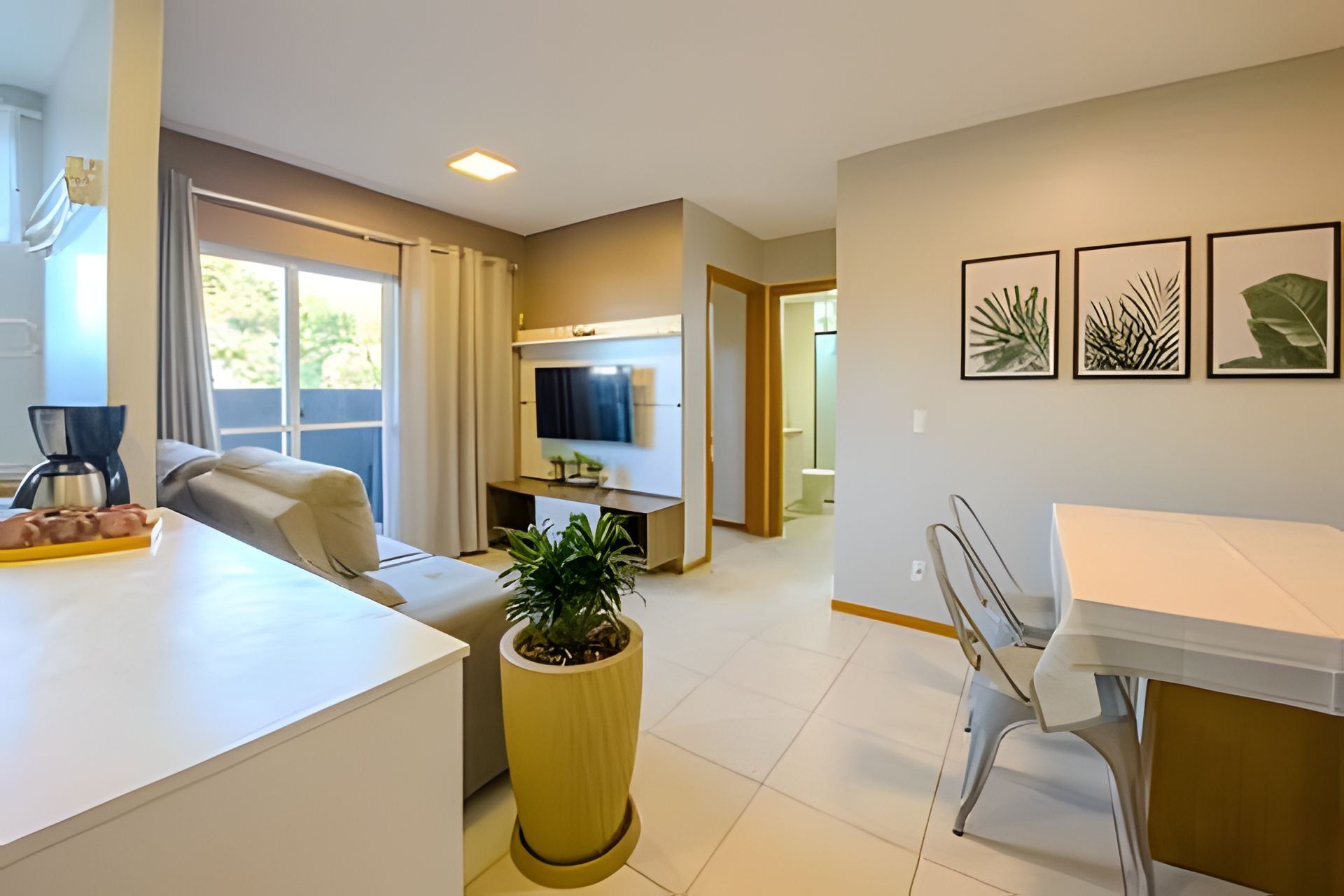 Una sala de estar moderna con un sofá, una mesa de centro y una planta en maceta. Se observa un televisor, un comedor con sillas y una cocina al fondo.
