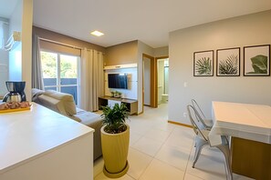 Una sala de estar moderna con un sofá, una mesa de centro y una planta en maceta. Se observa un televisor, un comedor con sillas y una cocina al fondo.