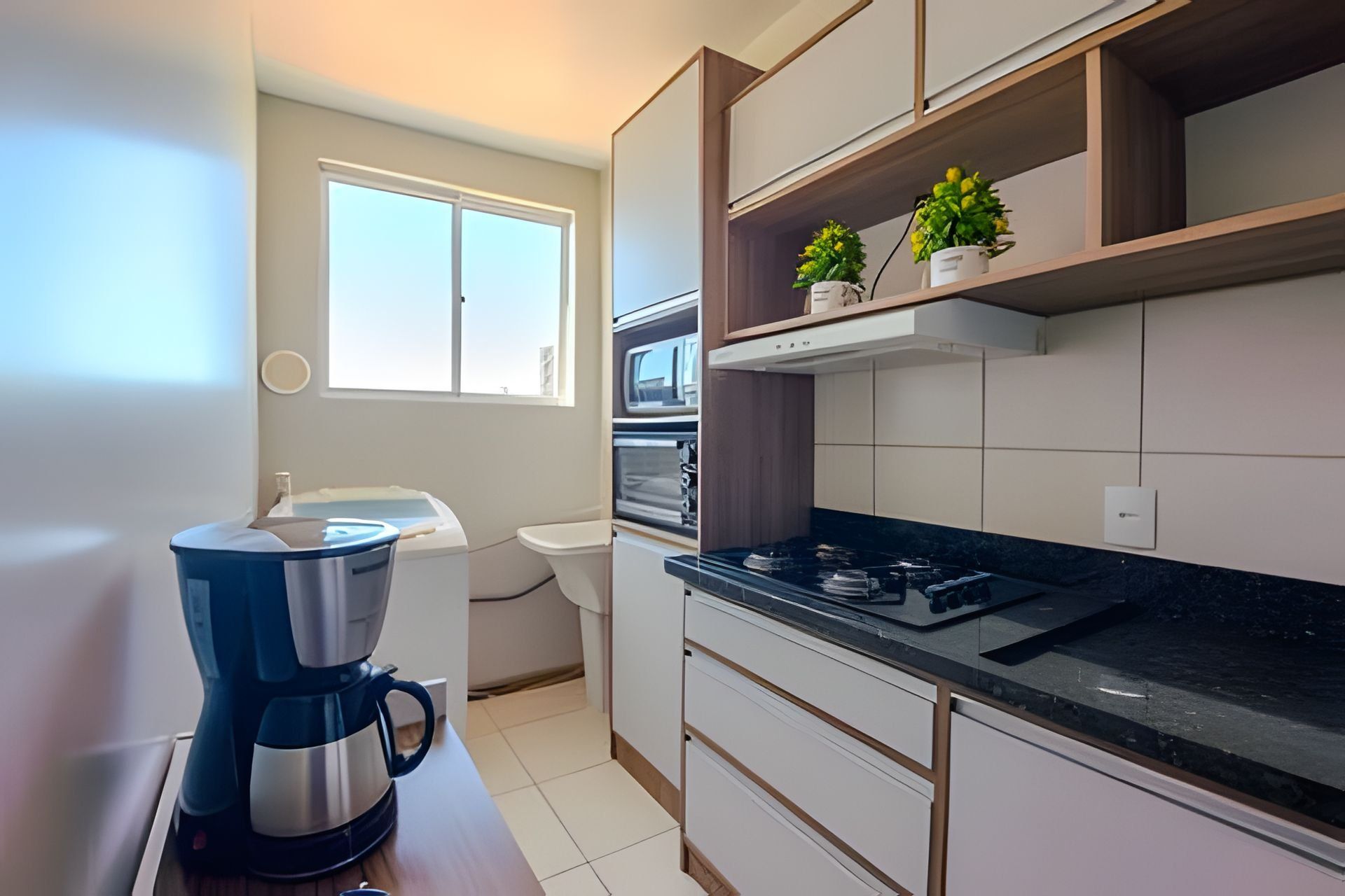 Cozinha compacta com cafeteira, micro-ondas e fogão. Há armários e uma janela.