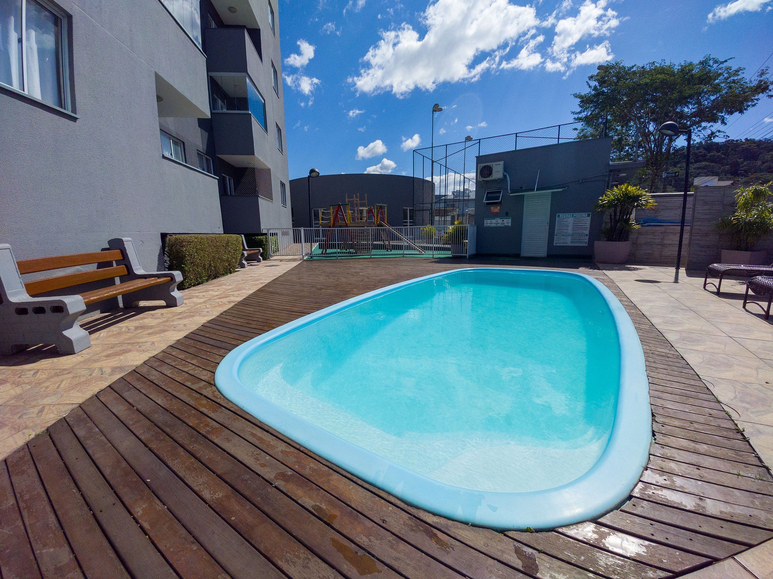 Une piscine extérieure avec une terrasse en bois, un banc et un bâtiment en arrière-plan.