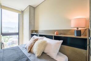 Ein Hotelzimmer mit einem großen Bett, einem Nachttisch, einer Lampe und Blick auf die Stadt durch ein Fenster.